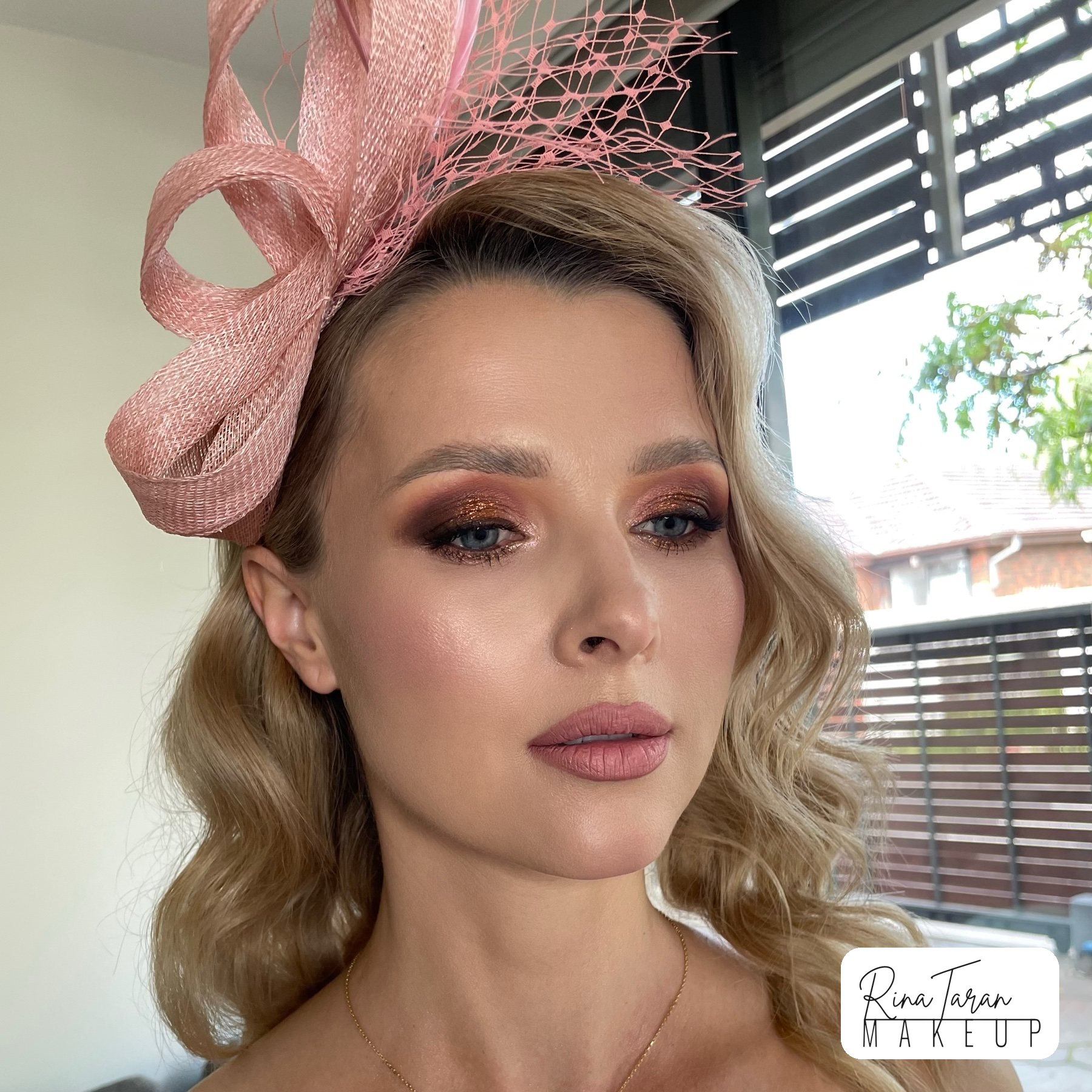 Race day makeup under a pink fascinator, warm copper eye and a glossy nude lip